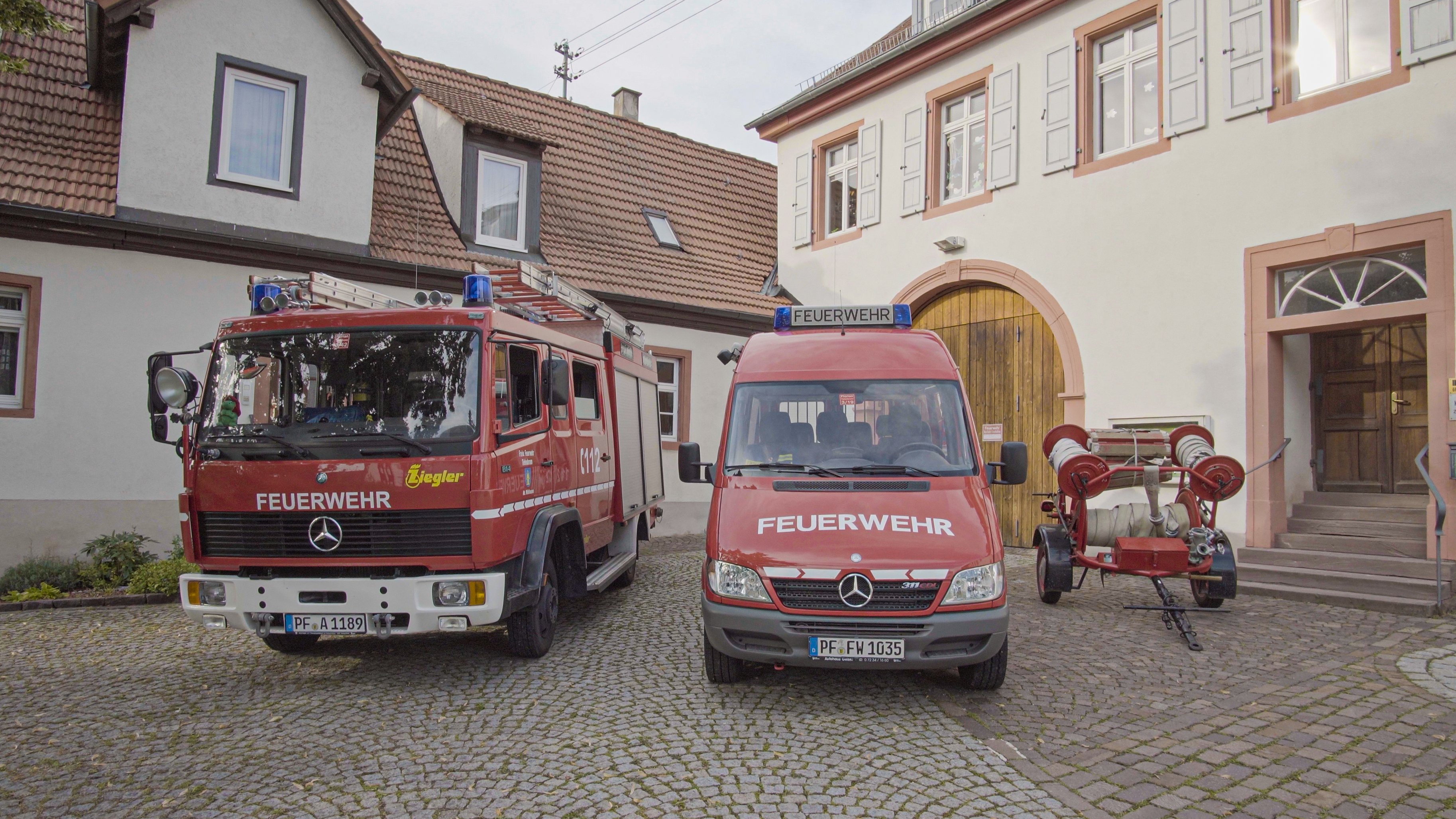 Zwei unterschiedliche Feuerwehrfahrzeuge und ein Schlauchanhänger von 1966 stehen vor dem Feuerwehrgerätehaus Mühlhausen, welches im Alten Schul- und Rathaus untergebracht ist.